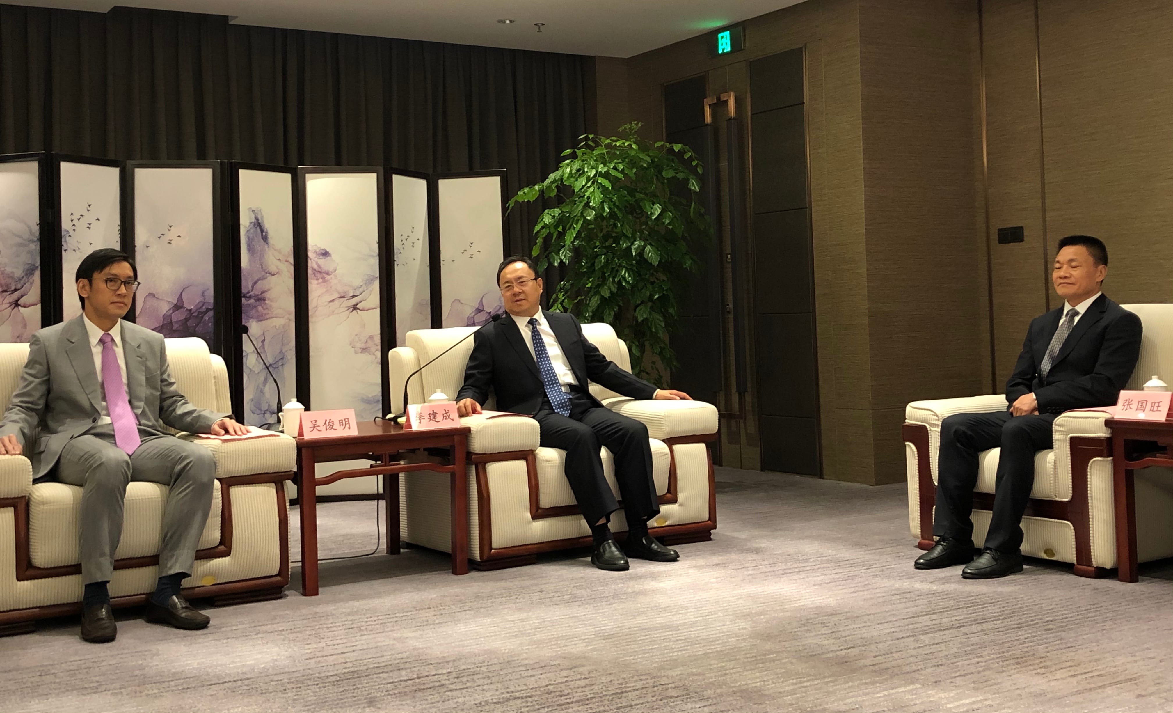 Three men in suits sit in armchairs in a conference room, nameplates on side tables.